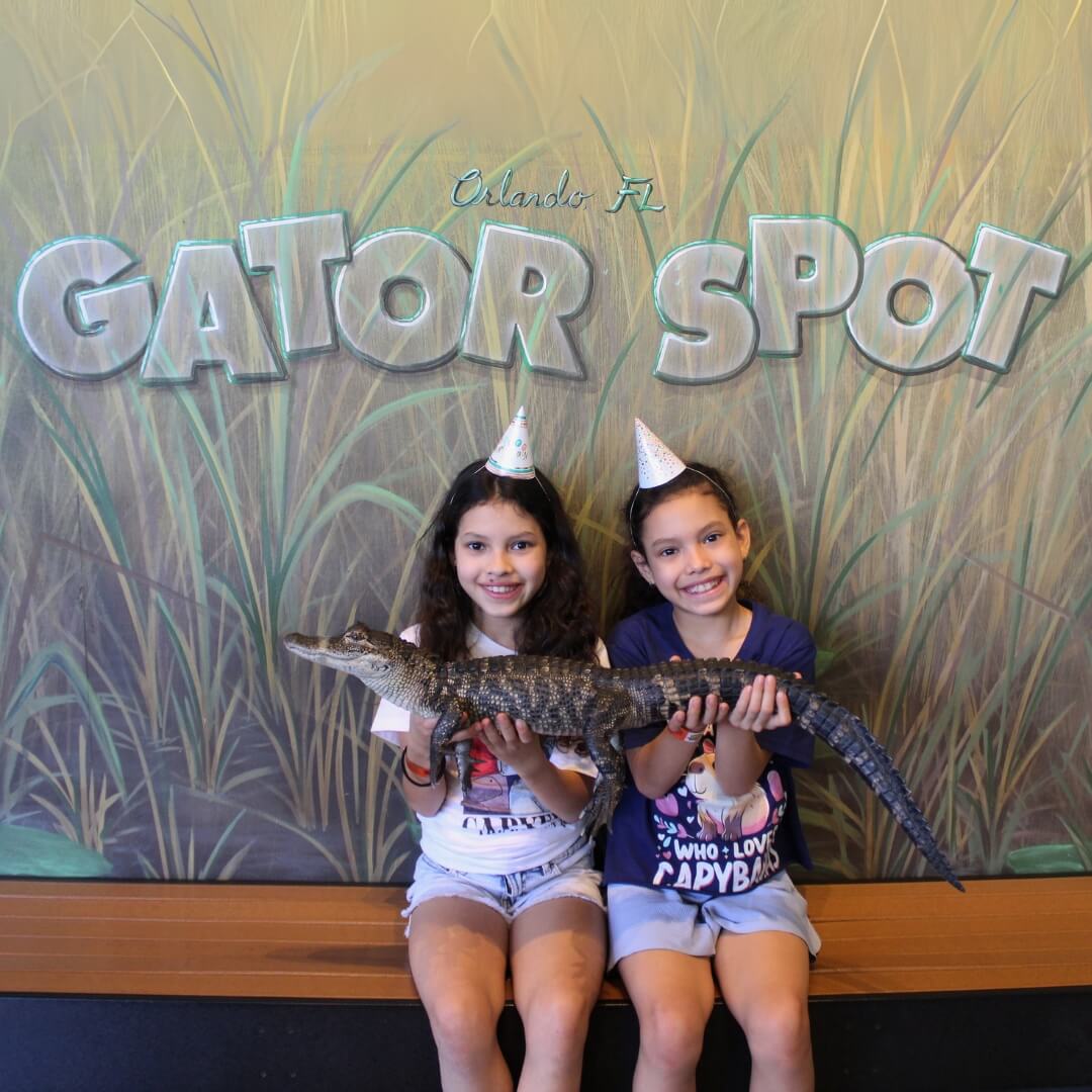 Two young girls wearing party hats hold a small alligator in front of a "GATOR SPOT" sign